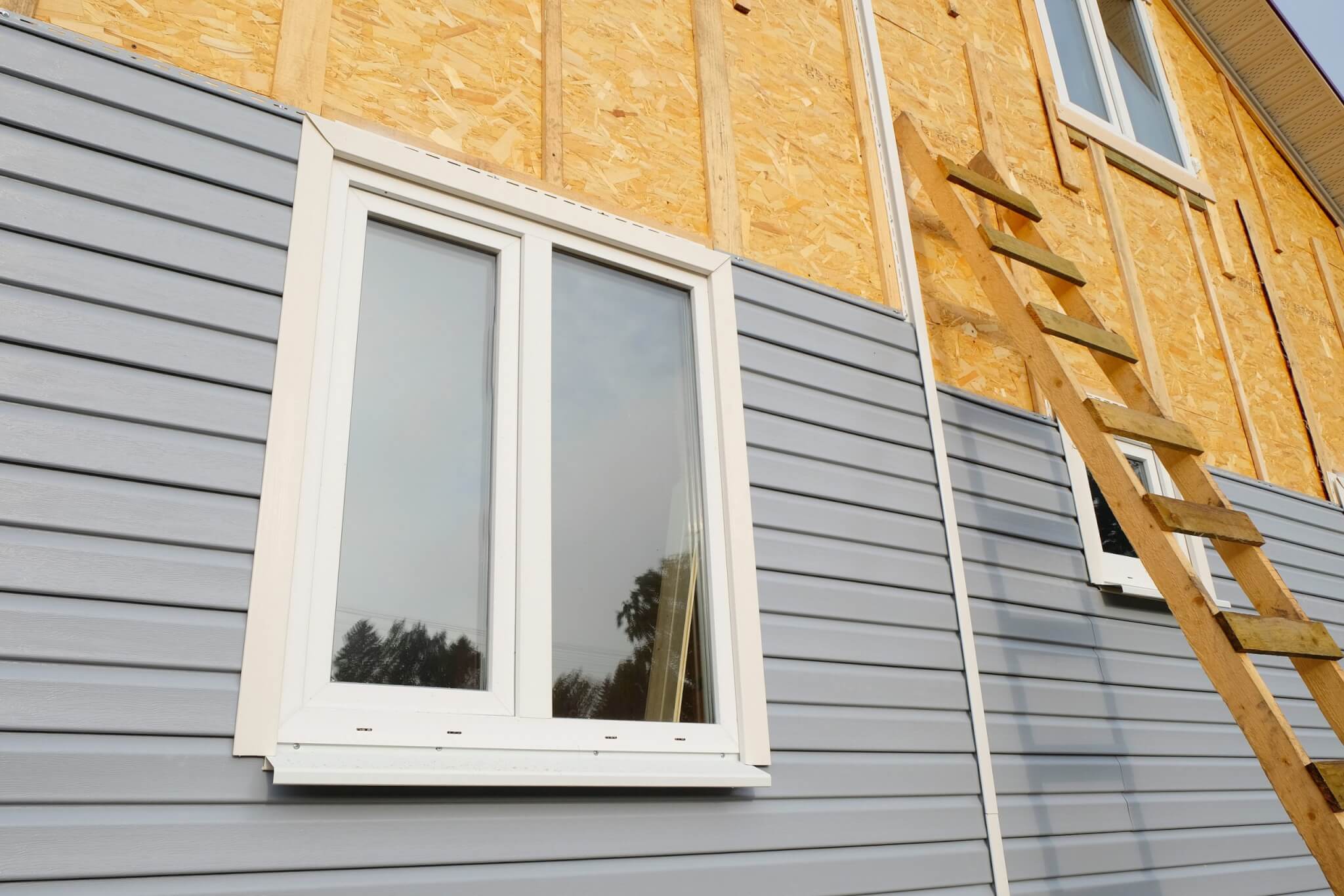 A charming house exterior features half wood paneling, half gray siding that showcases expert siding installation. A white window adds elegance, while a wooden ladder leans casually against the wall.