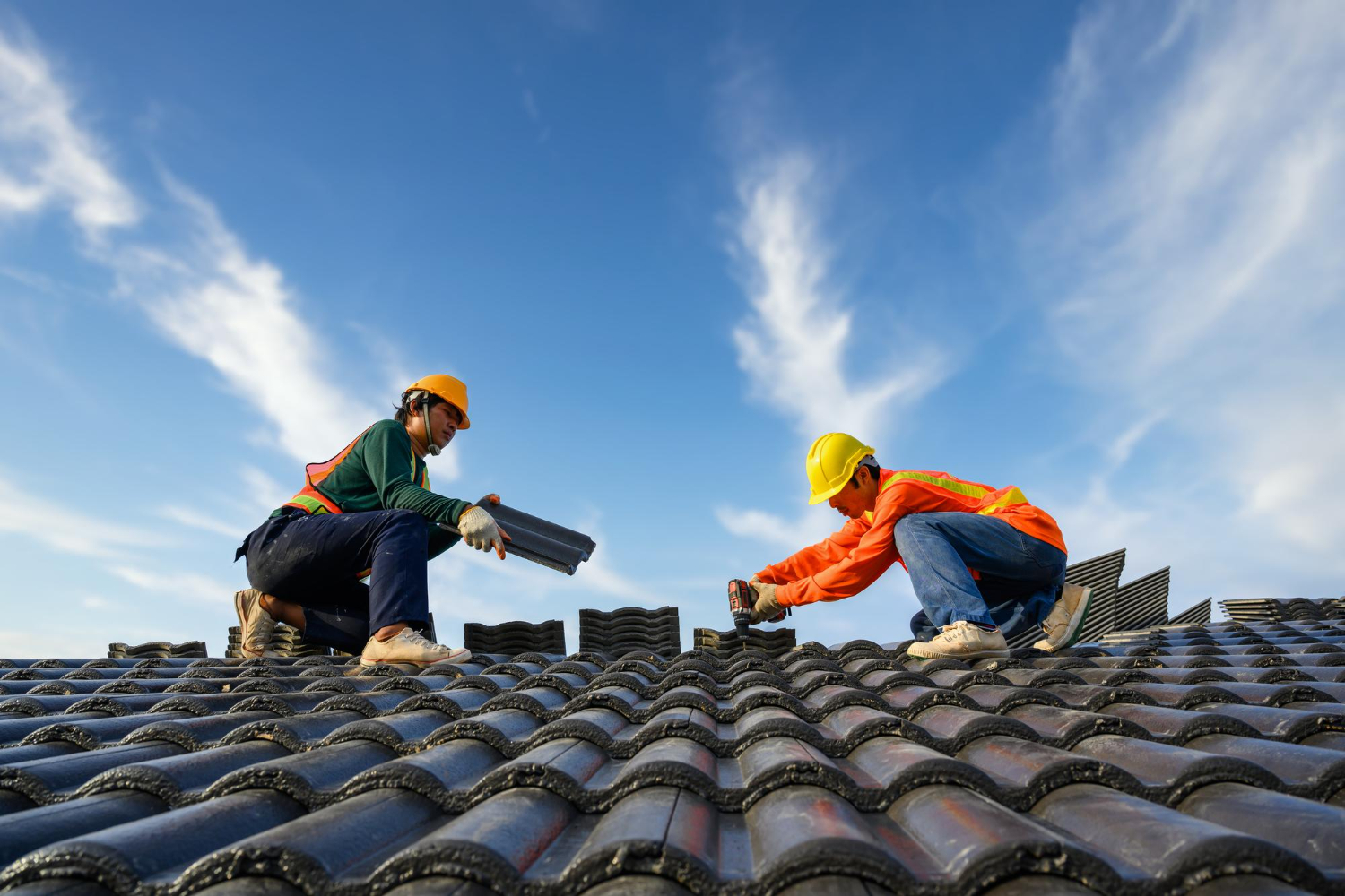 Two construction workers in safety gear installing roof tiles under a blue sky with clouds.