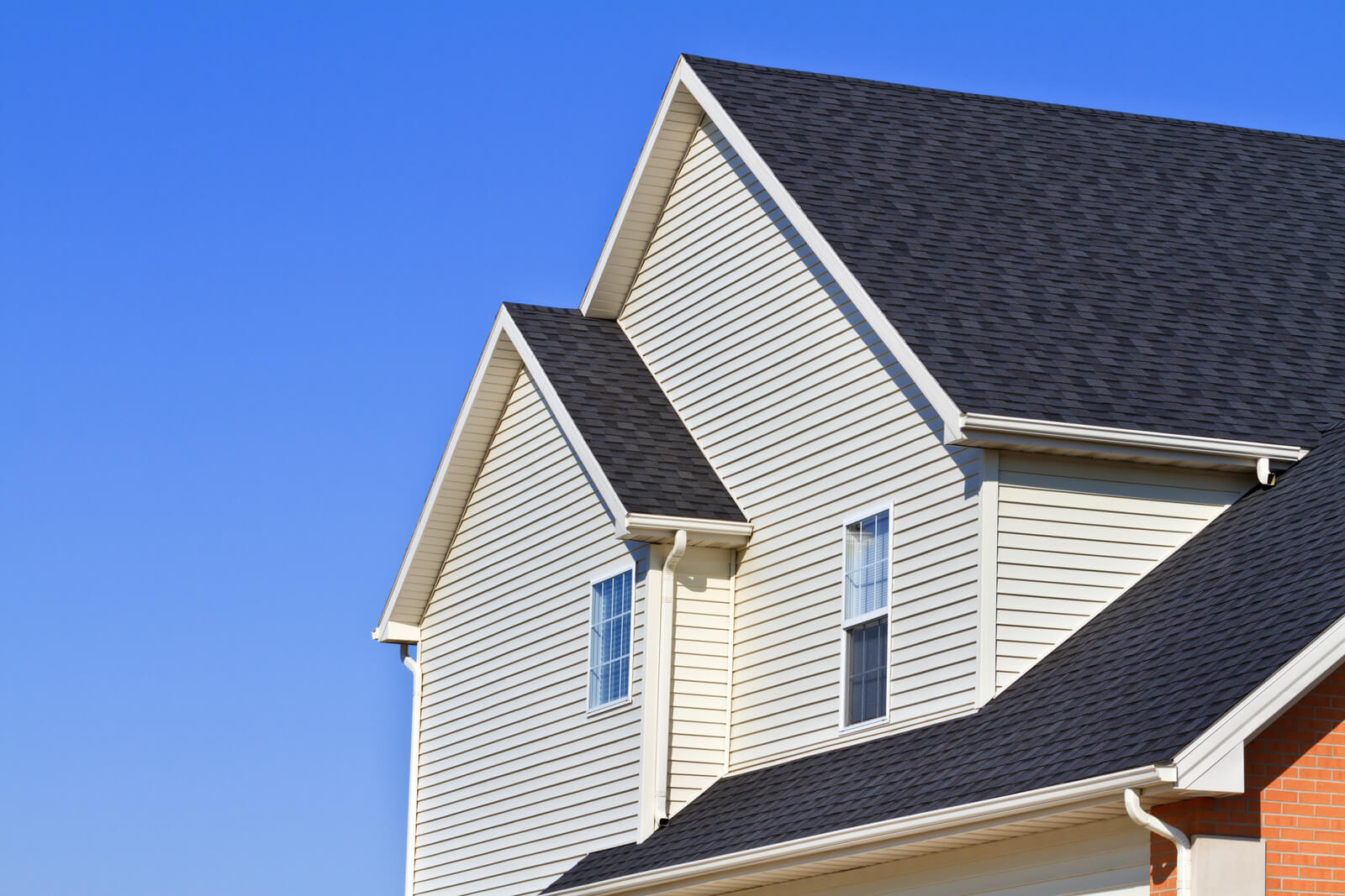 s2-5-1 Modern house with beige siding, black shingle roof, and white-trimmed windows against a clear blue sky.
